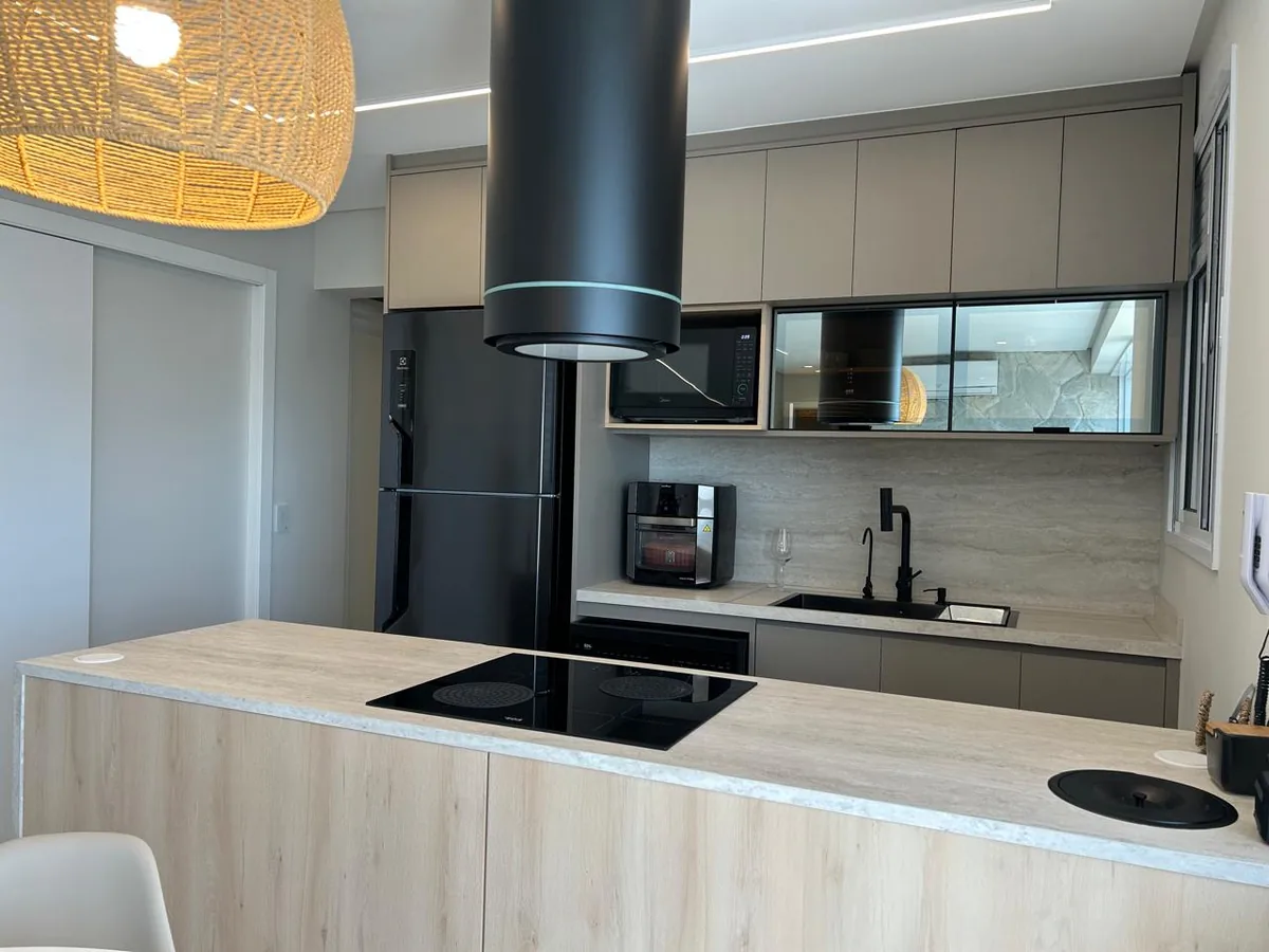 Cozinha com ilha de frente amadeirada e cooktop por indução, coifa cilíndrica preta, armários superiores em tom claro, nicho com micro-ondas, portas de vidro e pendente de palha