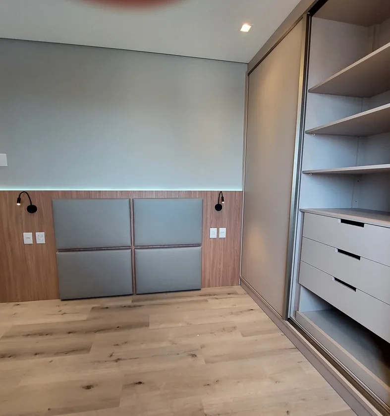 Dormitório com painel amadeirado, cabeceira estofada em módulos, arandelas e guarda-roupa de portas de correr com gaveteiro